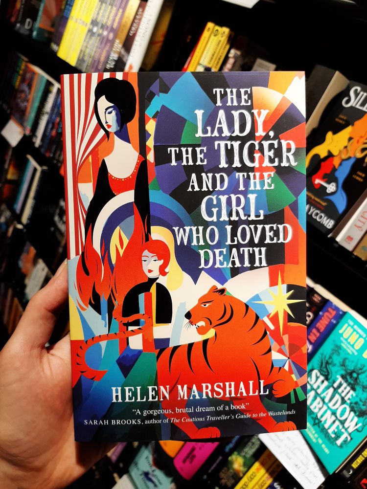 A hand holds a copy of the paperback book in a bookshop. The cover is bright and blocky in a circus kind if way, featuring a tiger, a figure whose red hair blends with flames in the background, and a black-haired woman, all in front of a colourful geometric and circus tent like set of patterns in the background.