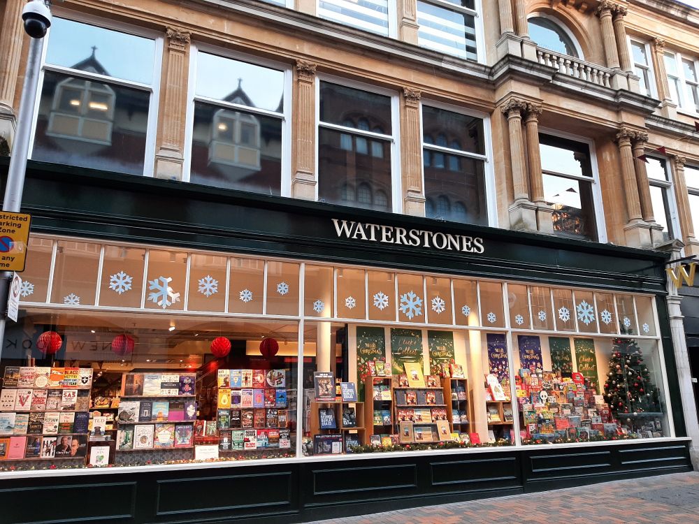 A golden glowing bookshop window decorated with paper snowflakes, a Christmas tree, lights, baubles and lots of wonderful books, games and gifts. The black windowframe features the word "Waterstones" in white.
