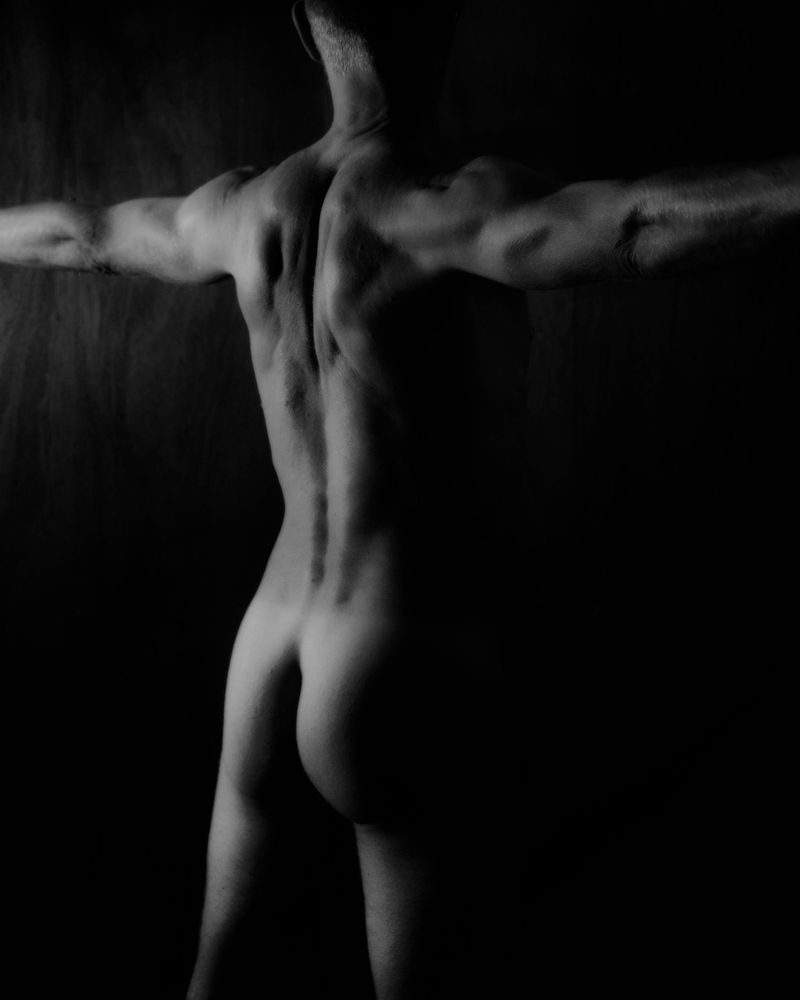 Nude man with his arms spread wide. Facing away from the camera so ass and back are visible. Single light source on the left creating a high contrast black and white image.