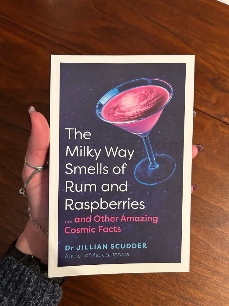 Front cover of the Milky Way Smells of Rum and Raspberries, held in the authors hand. A martini glass is filled with pink liquid with a galaxy swirl at the tip, against a starry dark blue background. 