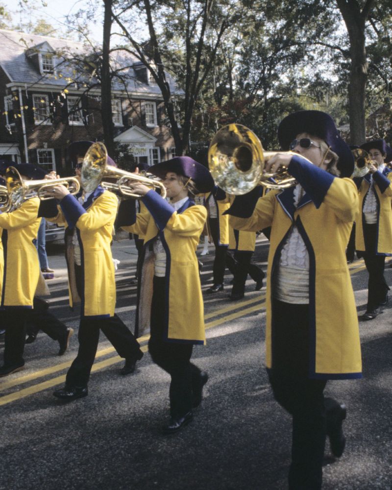 Members of a marching band perform in a parade, wearing yellow jackets and dark hats, playing trombones with traditional brick buildings in the background.