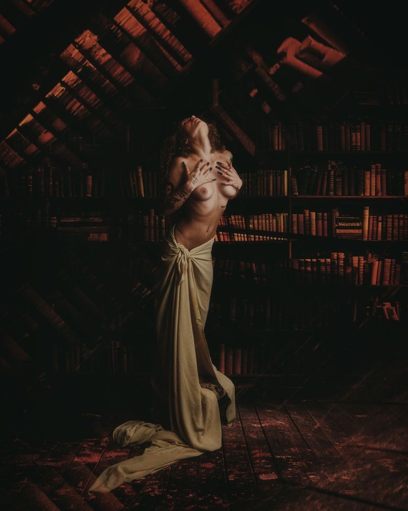 A woman stands elevated on a small platform, her legs draped in a robe that flows from her waist down, leaving her torso nude. Her head is tilted back and her hands rest gently on her chest. Behind her, bookshelves lined with old books fill a withered, decaying room. Her feet hover just above the floor, enhancing her ethereal, statue-like presence.