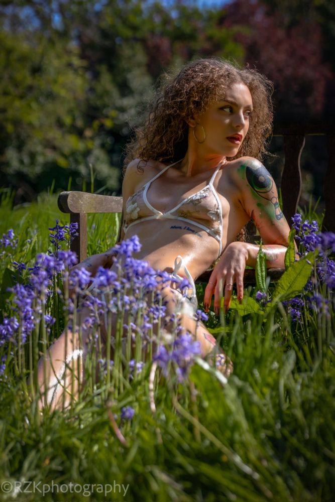 A woman sitting gracefully on a chair in the middle of tall green grass and blooming violet flowers, wearing delicate sheer lingerie, with natural light enhancing the ethereal outdoor setting.