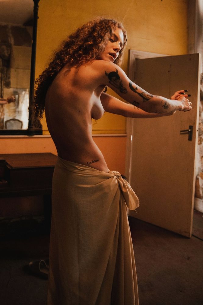 A woman stands confidently in a half-open robe, her torso nude, the robe is draping from the waist down against a backdrop of withered, surroundings. Her posture is statuesque, and she gazes cockily at the camera, evoking the presence of a defiant marble sculpture amidst ruins.