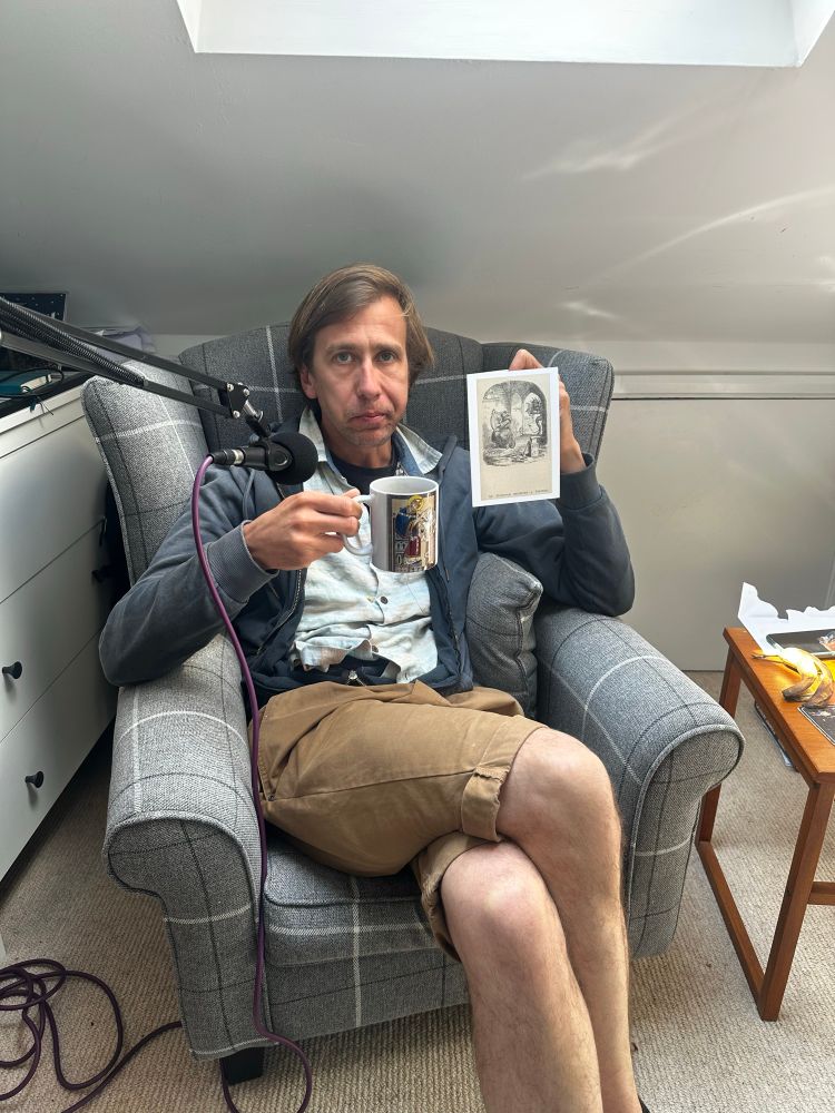 Ali sits on an armchair holding up a card with an image of the Saxon monk at Dunstable working in his forge as the devil enters, plus a mug with an image of Dunstable printed on it. Ali’s facial expression is grumpy.