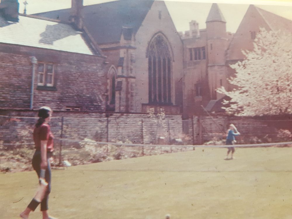 Two people play tennis on a grass court with the college in the background