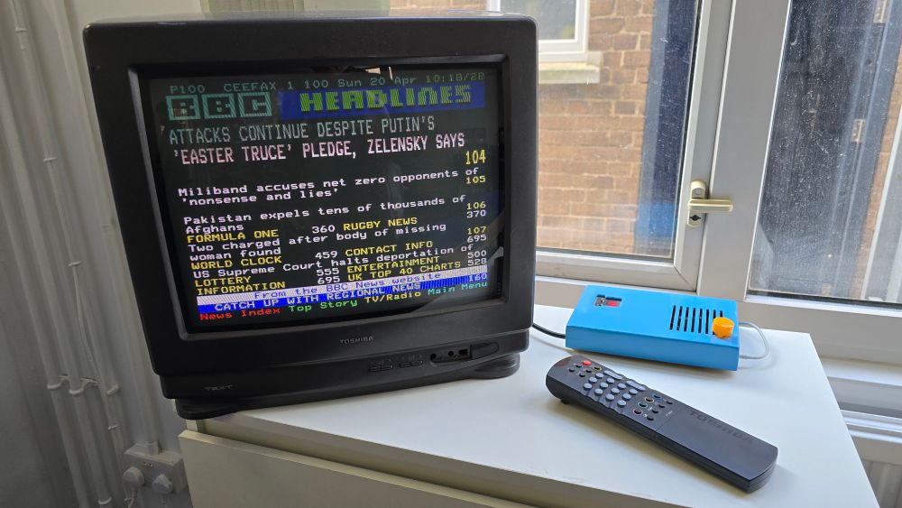 A retro CRT television displaying a BBC Ceefax teletext page with news headlines, sitting on a white table near a window. A blue teletext adapter box and a Toshiba remote control are also on the table.