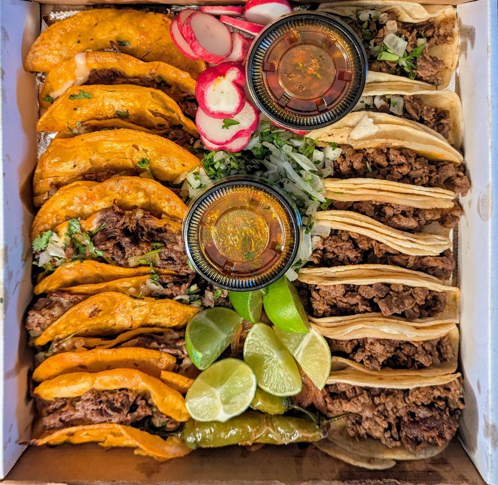 a box of 16 tacos half birria and half al pastor, birria consume, peppers, citrus, and veggies