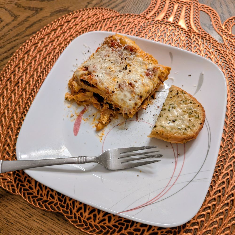 a crooked photo of a square of lasagna next to a heel of cheesy garlic bread on a white plate sitting atop a pumpkin shaped placemats on a wood table