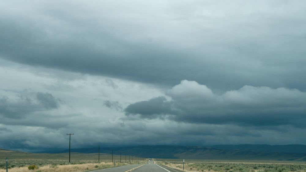 a straight road extends into the distance, and a horizontal row of dark hills and mountains, under a cloudy, stormy sky