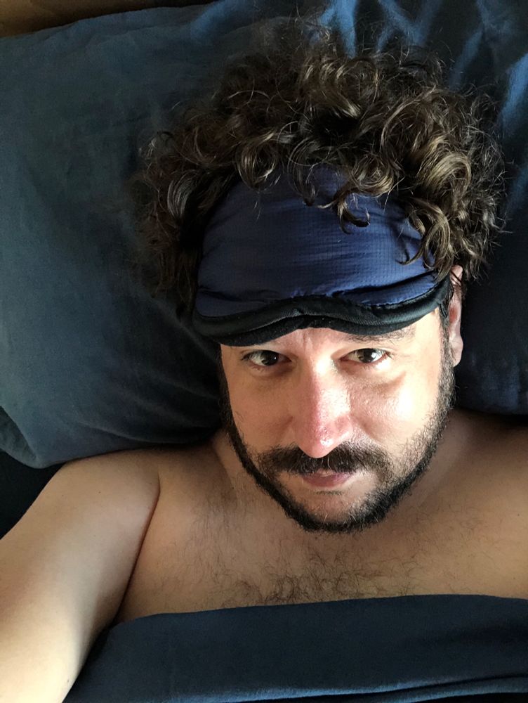 My face in bed against navy blue pillow with matching sheet covering most of my torso. Blue face mask on my forehead and curly brown hair out the top