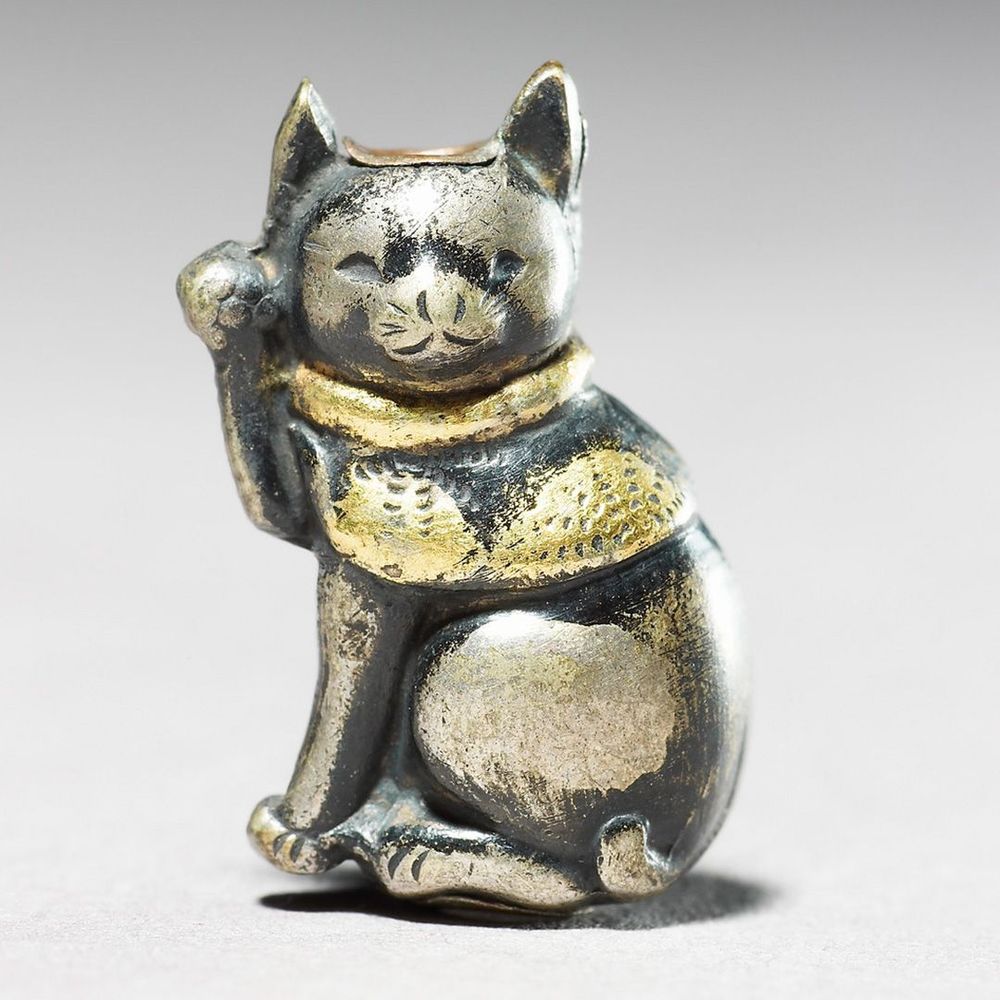 A silver metal ojime of a cat sat upright with a paw raised