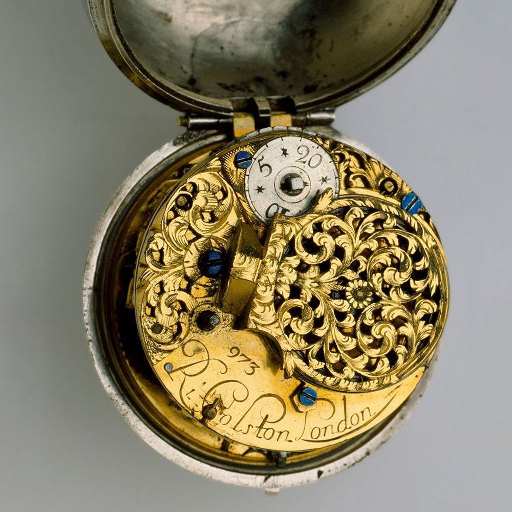 The inside workings of a round silver pocket watch. The inside mechanisms are hidden by a gold cover
