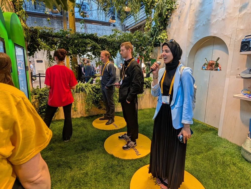 A person speaks into a microphone while others interact nearby in an indoor space with artificial grass, plants, and decorative elements.