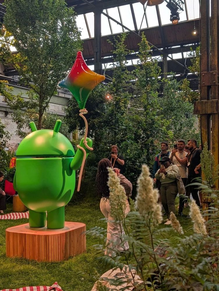 A green Android mascot statue holds a star-shaped balloon in a garden-like indoor space. Attendees take photos nearby.