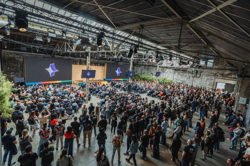 A large audience sits and stands in a spacious industrial room with high ceilings and exposed walls, facing a stage. 