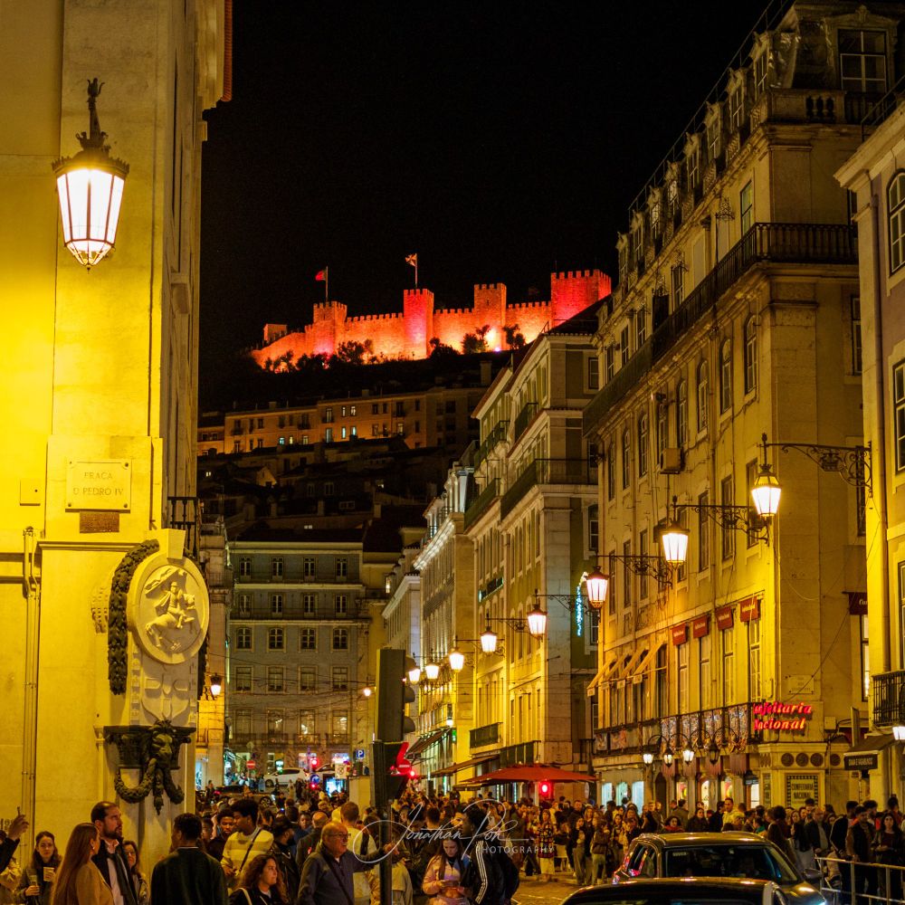 A street scene in Lisbon at Christmas time, crowds of people, traditional lamps hanging on the buildings. The São Jorge Castle (Castelo de São Jorge) is visible in the background between the buildings, illuminated with red lights.