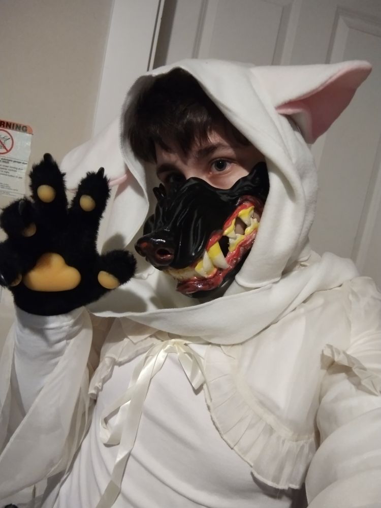 @owlhops in lacy white clothing, with a hood that has lamb or sheep like ears. Beneath the hood, over their nose and mouth, is a snarling black wolf mask with yellowed teeth and red gums. they wear black fursuit paws, one raised up to show the pawpads and claws.