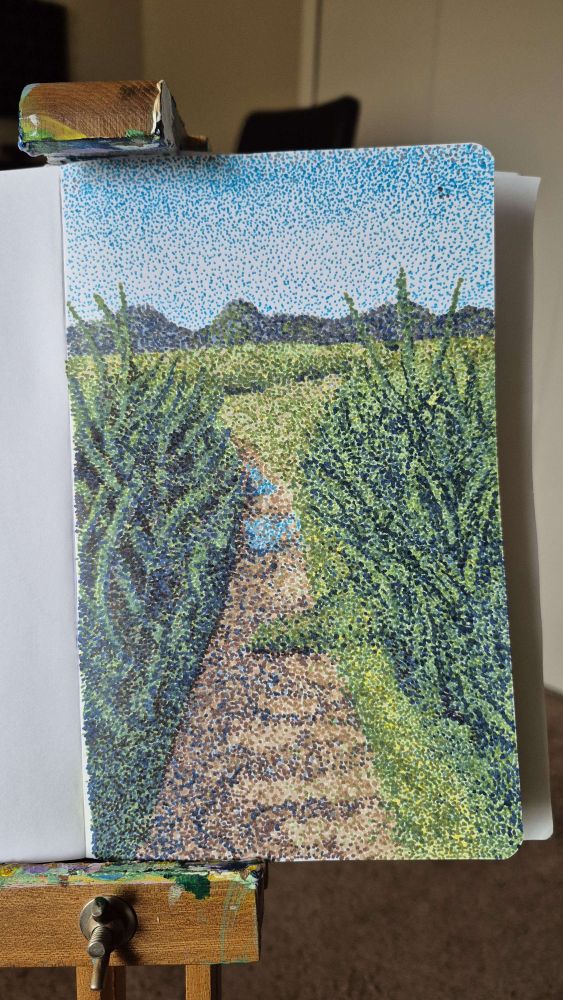 A sketchbook on an easel open to a page showing a pointillism landscape of a path through reeds. In the distance of the landscape are mountains and a blue sky.