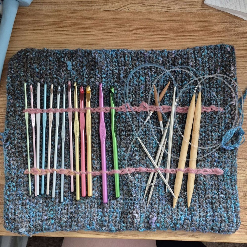 A blue and grey crocheted case. It is open and various crochet hooks and circular knitting needles are held in play by pink surface crocheted lines.