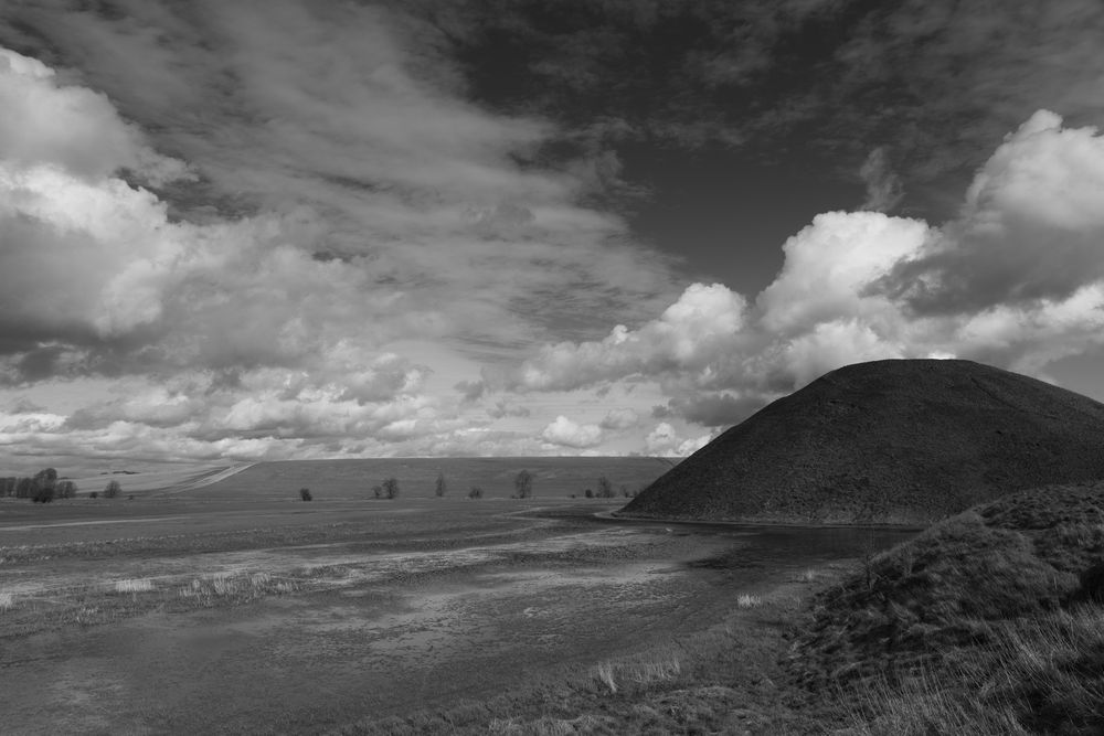 A black and white landscape  photo of Silbury Hill, a large neolithic earthen mound, in context in the Wiltshire landscape.