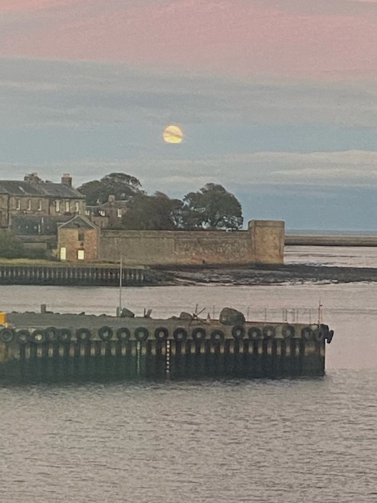 Huge harvest moon rising across the Tweed estuary at Berwick. Sky is grey blue and pink reflected in the river water
