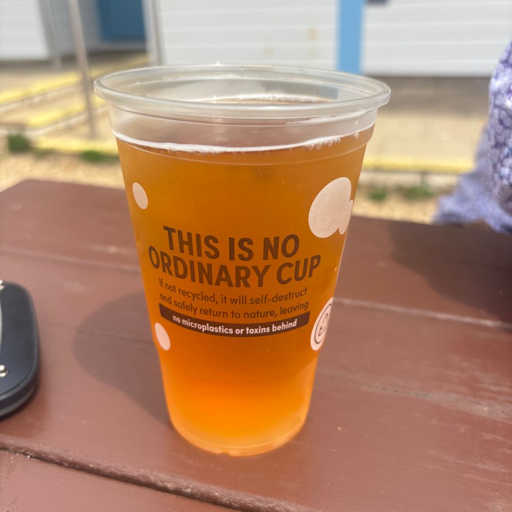 Plastic pint cup full of beer on a wooden outdoor table. On the cup it says “THIS IS NO ORDINARY CUP If not recycled, it will self-destruct and safely return to nature, leaving
no microplastics or toxins behind”