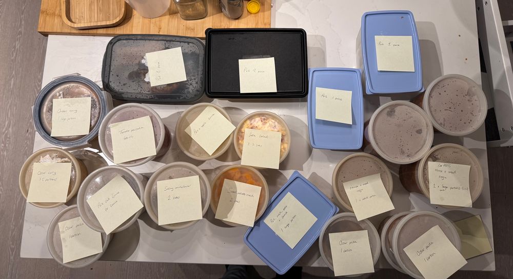 A variety of containers filled with different sorts of food are lying on top of a kitchen table. The containers have post-it notes on them describing their contents