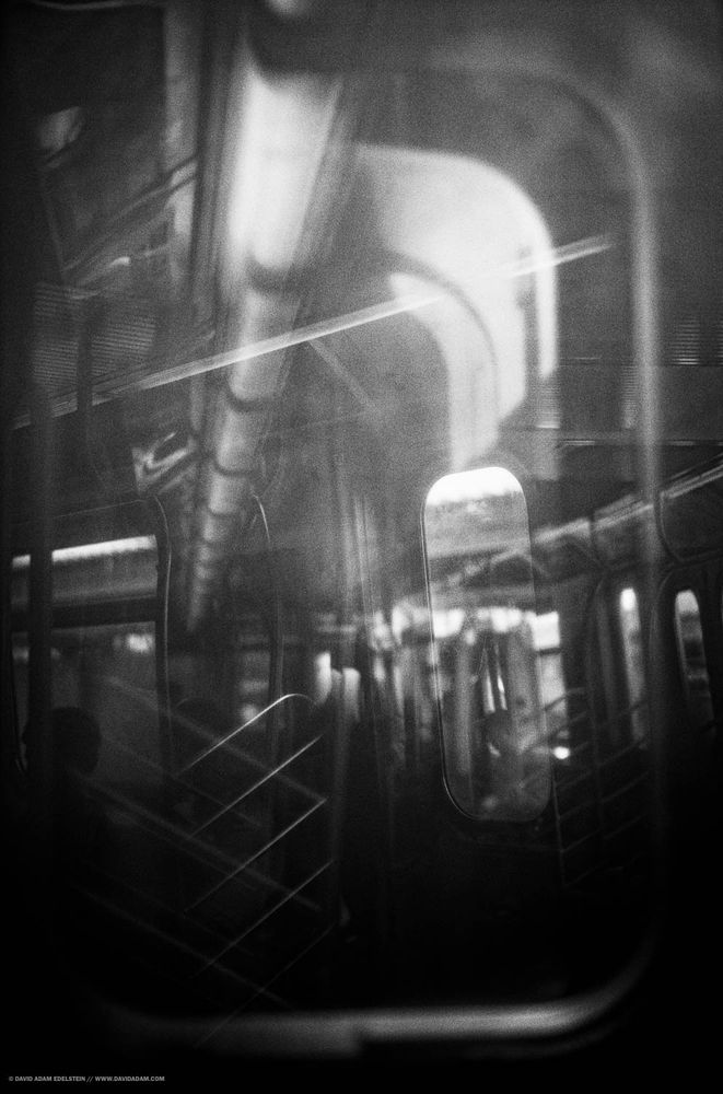 Black and white photo looking from one subway car into the next. The layered reflections between the cars make it hard to tell what is where.