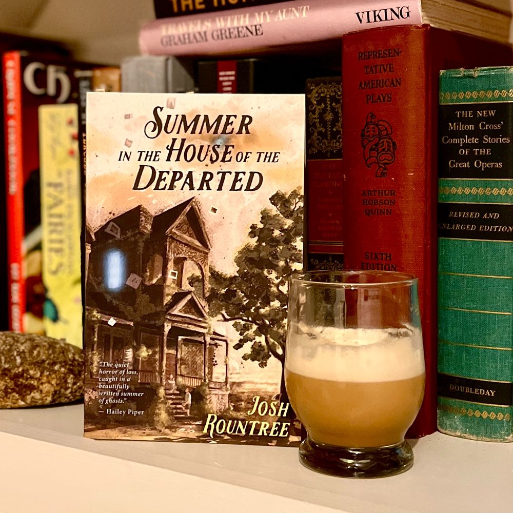 SUMMER IN THE HOUSE OF THE DEPARTED by Josh Rountree, a coffee colored book alongside a coffee drink on a bookshelf.