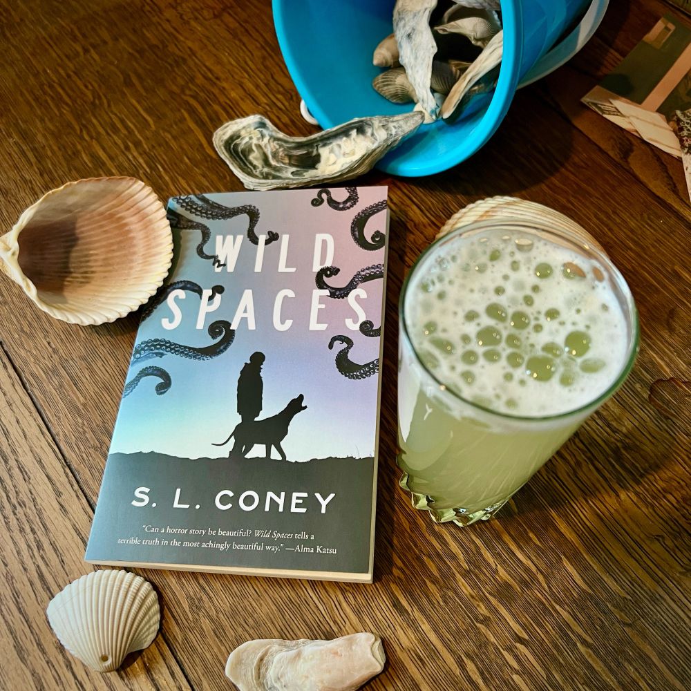 [image desc: WILD SPACES by S.L. Coney on a wooden surface surrounded by shells and a tall pale yellow-green highball.]