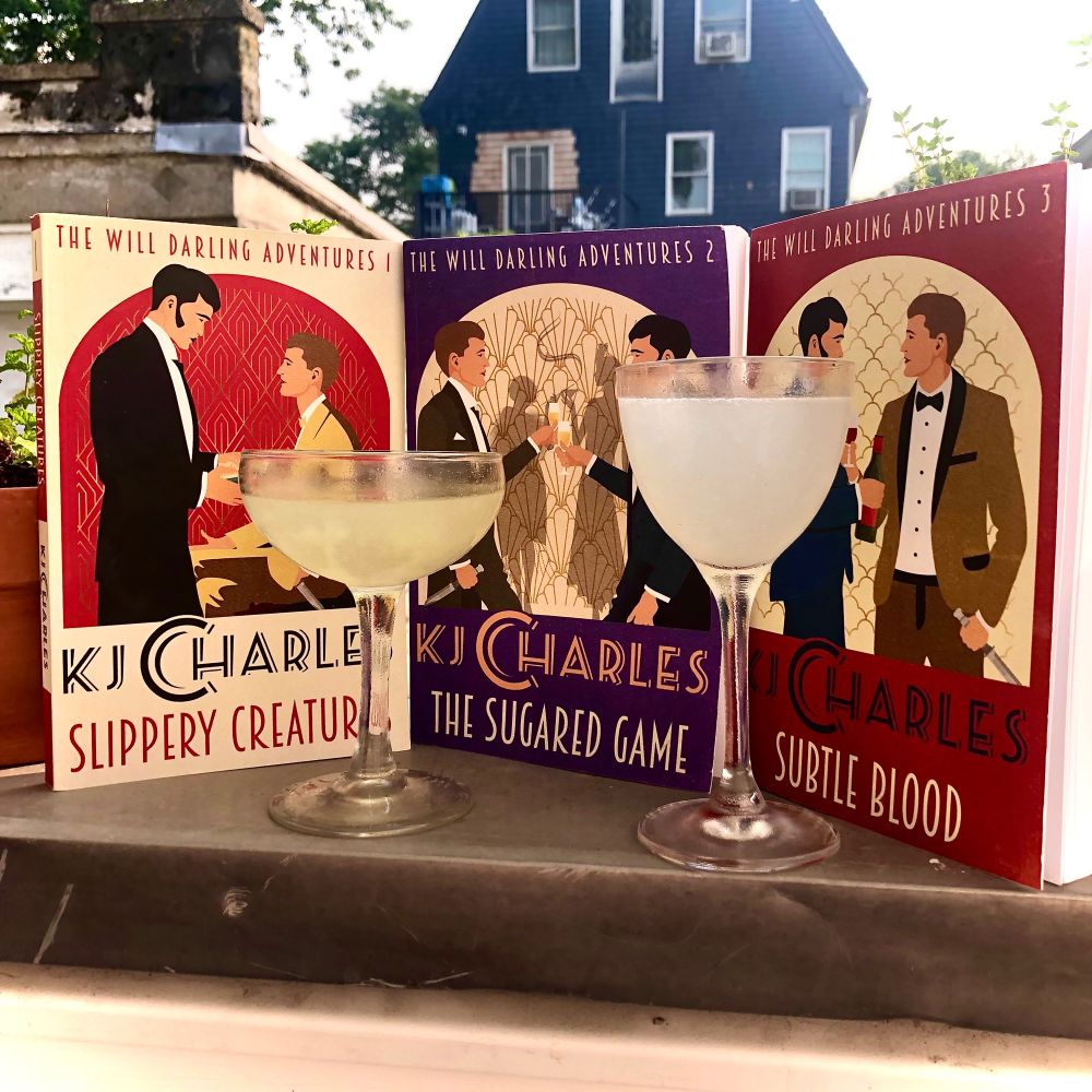 Two glasses: with a rich green and pale green gimlet sit between the three books in KJ Charles’ WILL DARLING ADVENTURES trilogy of historical romances.