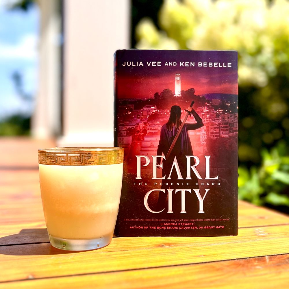 [A pale drink in a gold rimmed glass on a cedar porch alongside PEARL CITY by Julia Vee and Ken Bebelle.]