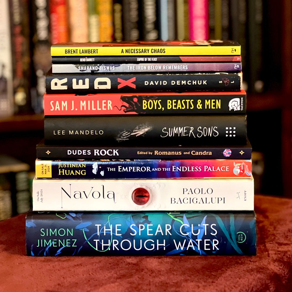 A stack of books (bottom to top): THE SPEAR CUTS THROUGH WATER by Simon Jimenez; NAVOLA by Paolo Bacigalupi; THE EMPEROR AND THE ENDLESS PALACE by Justinian Huang; DUDES ROCK Ed. Romanus and Candra; SUMMER SONS by Lee Mandelo; BOYS, BEASTS & MEN by Sam J. Miller, RED X by David Demchuk; THE IRON BELOW REMEMBERS by Sharang Biswas; EMPIRE OF THE FEAST by Bendi Barrett; A NECESSARY CHAOS by Brent C. Lambert