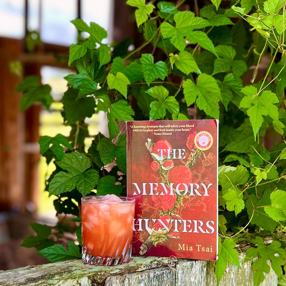 A red drink in a low glass alongside an advance copy of THE MEMORY HUNTERS by Mia Tsai. The book and drink are on a weatherbeaten wooden rail, flanked by greenery and slightly backlit by a far wall missing paneling.