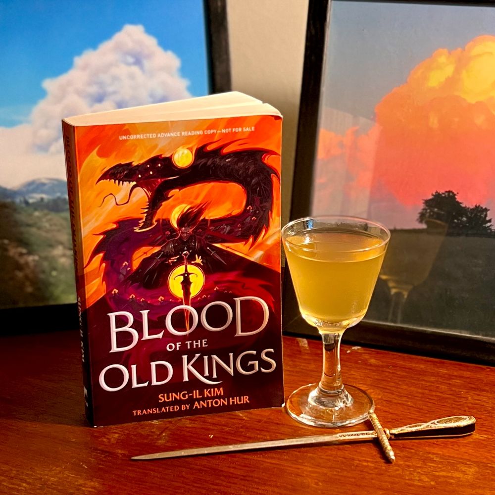 A copy of BLOOD OF THE OLD KINGS by Sung-il Kim (translated by Anton Hur) sits on a wooden surface alongside a pale yellow-gold drink in a stemmed glass. A letter opener like a sword sits before the book and drink and two framed photos of white smoke and golden cloud sit in the background