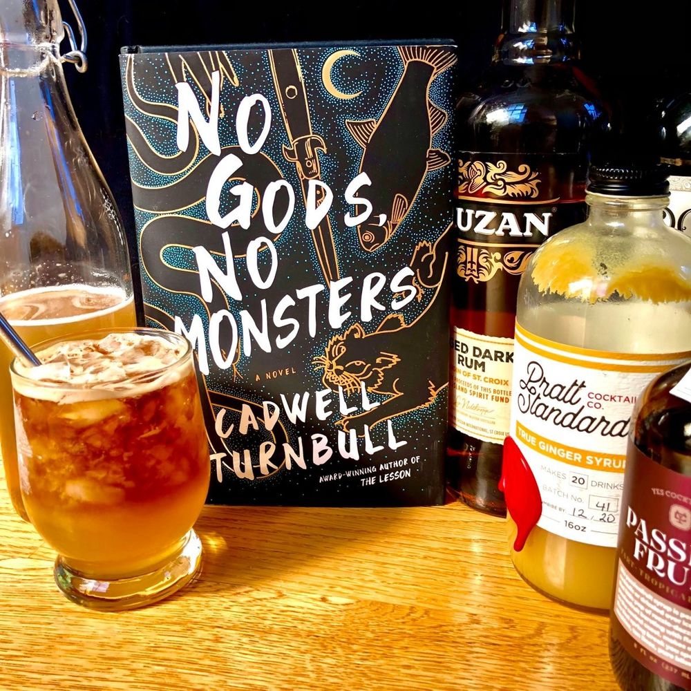 NO GODS, NO MONSTERS by Cadwell Turnbull is flanked by a low glass filled with crushed ice and a layered cocktail in gold and black. On the other side is a bottle of Cruzan aged rum, Pratt Standard ginger syrup, and Yes! Cocktail Co. Passion Fruit Syrup.
