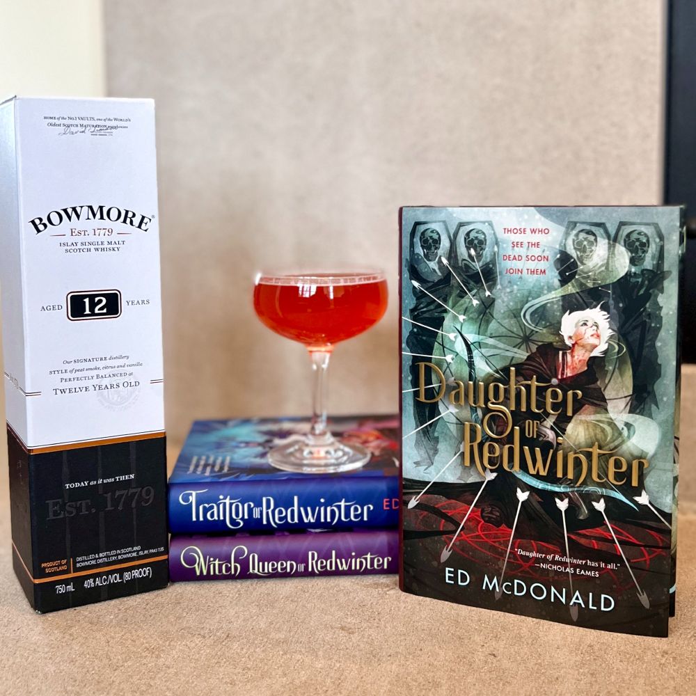 A bright red cocktail sits atop TRAITOR OF REDWINTER and WITCH QUEEN OF REDWINTER by Ed McDonald. They are flanked by a box containing a bottle of the Bowmore 12 yr scotch whisky and DAUGHTER OF REDWINTER by Ed McDonald.