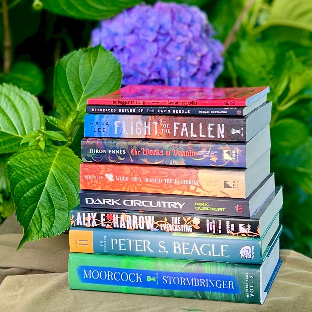 A stack of books in front of a deep blue-purple hydrangea (bottom to top): STORMBRINGER (THE ELRIC SAGA vol. 2) by Michael Moorcock, THE BEST OF PETER S. BEAGLE vol. 2 by Peter S. Beagle, THE EVERLASTING by Alix E. Harrow, DARK CIRCUITRY by Kirk Bueckert, WHEN THEY BURNED THE BUTTERFLY by Wen-yi Lee, THE WORKS OF VERMIN by Hiron Ennes, FLIGHT OF THE FALLEN by Hana Lee, DISGRACED RETURN OF THE KAP’S NEEDLE by Renan Bernardo, THE DAGGER IN VICHY by Alastair Reynolds