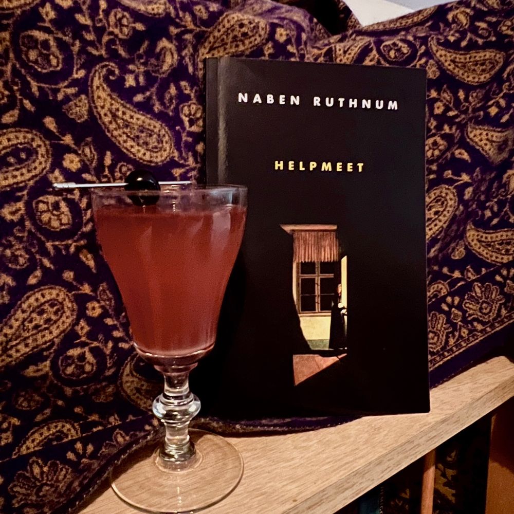 [image desc: A dark reddish mocktail garnished with a cocktail cherry sits beside a copy of HELPMEET by Naben Ruthnum. The mood is dark and atmospheric.]