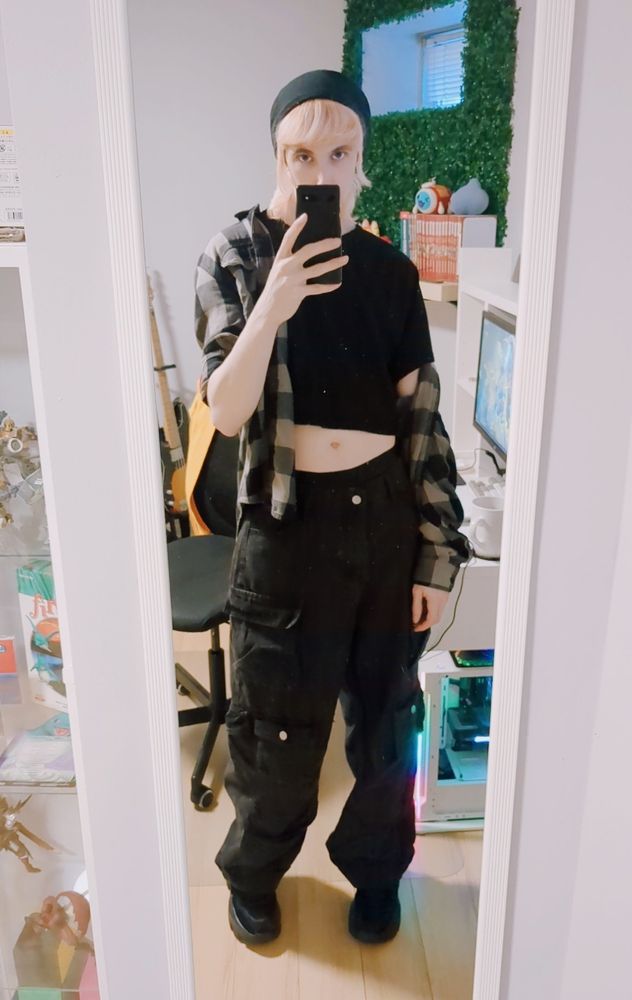 a selfie of mlemmy! hes wearing baggy jeans, chunky sneakers, a crop top and a flannel over top, and a beanie!!