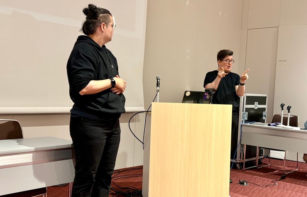 Two people presenting on opposite ends of a podium. On the left, the deaf presenter (Oliver) looks on while their hearing collaborator (Katta) is signing on the right. 