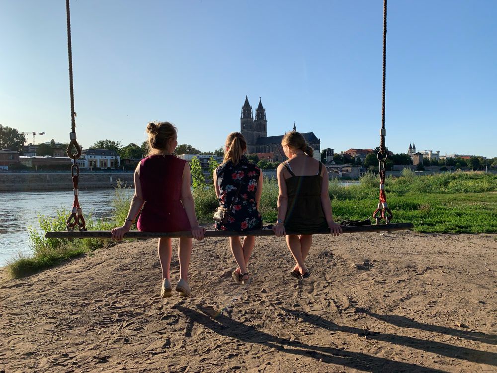 Im Licht der tief stehenden Sonne sitzen drei Frauen auf  einer gut drei Meter breiten Schaukel, den Blick auf die Elbe und die Altstadt Magdeburgs im Hintergrund gerichtet.