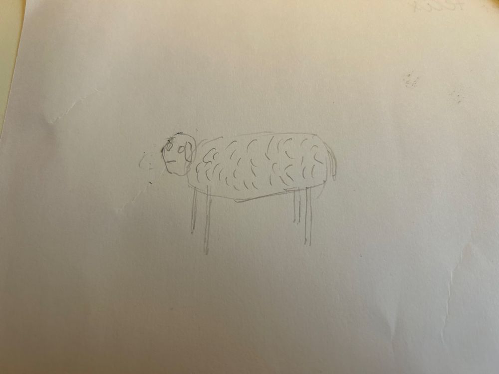 A pencil drawing on paper of a sheep drawn from memory. It looks not very realistic consisting of a large, oval body with some „waves“ indicating its wooly coat. It has a head with two circles for eyes and two ears and a mouth without further details. Legs are placed like you’d expect them to for a standing sheep but also lack any further detail. A tail is hinted at. 