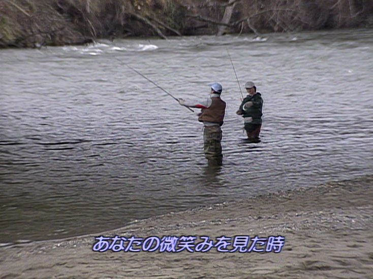 Two men fishing in a river with a Japanese subtitle reading (italicized) "あなたの微笑みを見た時"