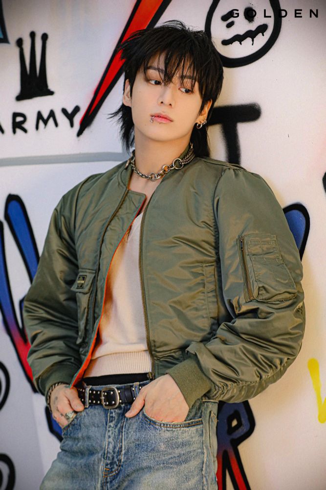 Jungkook of BTS standing with hands in pockets leaning against a wall with graffiti on it. Wearing green jacket and jeans with a pensive expression. 