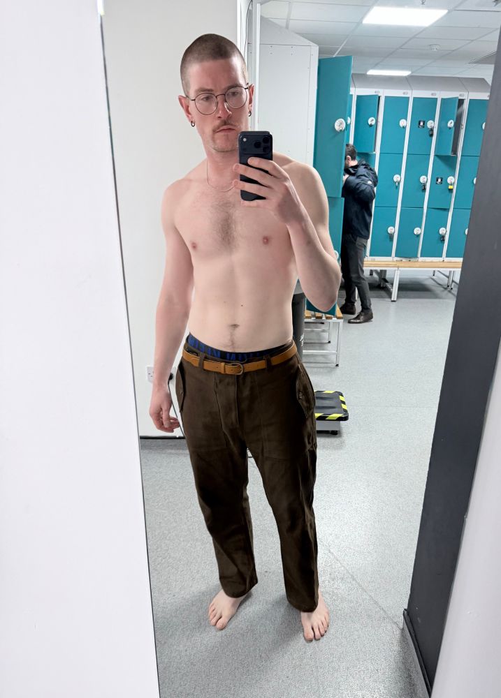 A man in a gym changing room wearing brown trousers but topless and shoeless