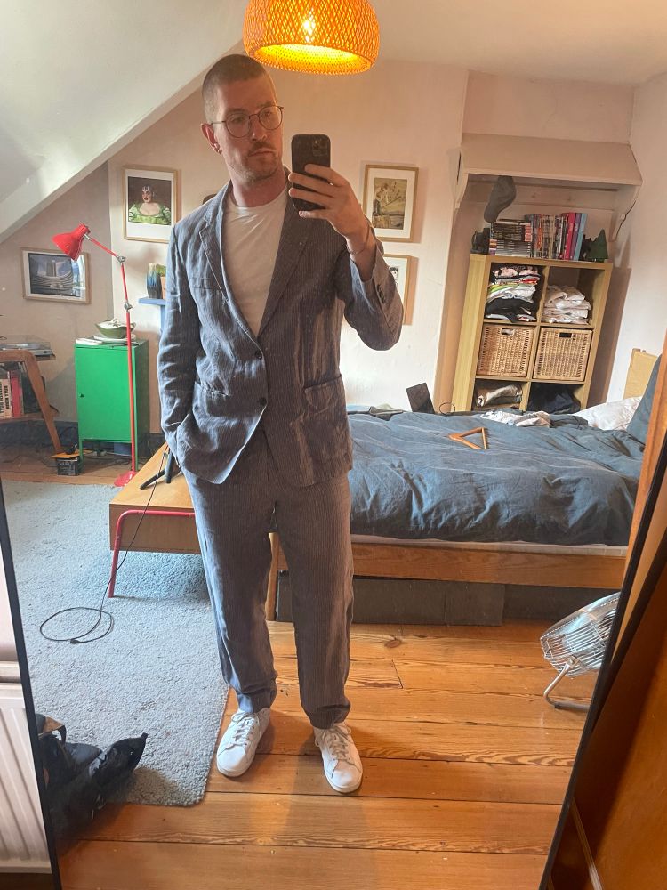 A man stands in a blue striped linen suit with a white T-shirt and trainers