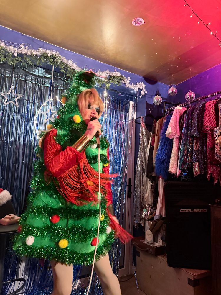 A drag queen in a Christmas tree outfit holding a mic with racks of clothes behind them