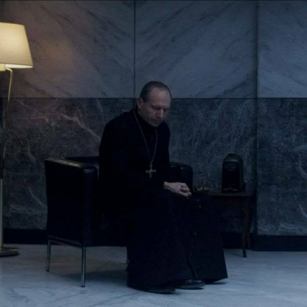 Ralph Fiennes as the archbishop in the movie Conclave. He is sitting down and praying, looking beleaguered 